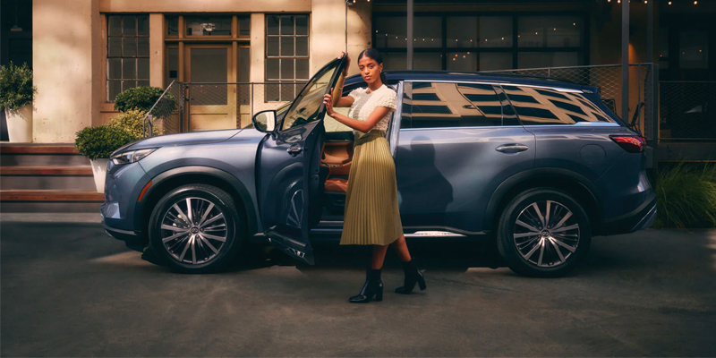 A woman in a yellow skirt standing next to a blue 2025 INFINITI QX60 SUV with its door open.